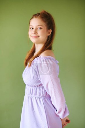 Photo for Beauty portrait of pretty young 15 - 16 year old redhaired teeenage girl wearing purple dress posing on green background - Royalty Free Image