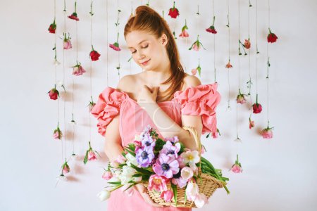 Téléchargez les photos : Portrait studio de jolie jeune adolescente de 15 à 16 ans rousse portant une robe de corail rose, posant sur fond blanc avec des roses suspendues, tenant panier avec de nombreuses fleurs de printemps, concept de beauté et de mode - en image libre de droit