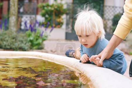 Outdoor portrait of adorable little boy playing outside, mother stopping kid from getting into old dirty fountain