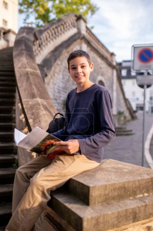 Téléchargez les photos : Joyeux adolescent assis sur les escaliers en béton balustrade avec un livre ouvert regardant devant lui - en image libre de droit