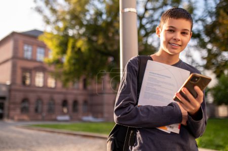Foto de Retrato en la cintura de un niño alegre con el teléfono celular y el libro de texto mirando delante de él - Imagen libre de derechos