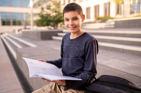 Cheerful dark-haired teenage boy sitting on the porch of a building with an open textbook