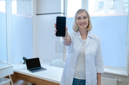 Téléchargez les photos : Femme médecin souriante et joyeuse en blouse blanche tenant son smartphone devant la caméra - en image libre de droit