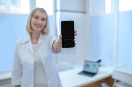 Téléchargez les photos : Agréable femme médecin vêtue de la blouse blanche tenant son téléphone portable devant la caméra - en image libre de droit