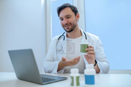 Foto de Retrato de cintura hacia arriba de un médico sosteniendo una botella de vitamina plástica antes de la pantalla del portátil durante la consulta de vídeo - Imagen libre de derechos