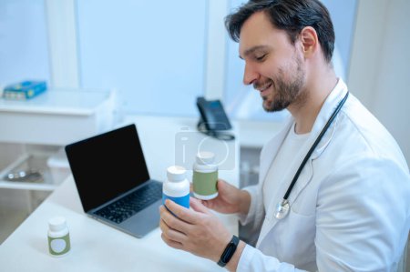 Téléchargez les photos : Médecin souriant assis au bureau avant l'ordinateur portable regardant les bouteilles de vitamines en plastique dans ses mains - en image libre de droit
