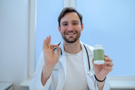 Foto de Retrato en la cintura de un médico amigo sonriente con la botella de vitamina plástica en la mano haciendo el signo OK - Imagen libre de derechos