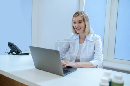 Téléchargez les photos : Médecin concentrée souriante dans la blouse blanche tapant sur l'ordinateur portable dans son bureau - en image libre de droit