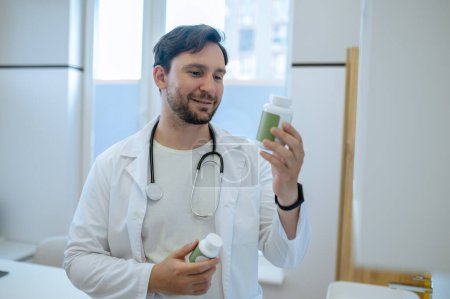 Foto de Retrato en la cintura de un médico sonriente enfocado en una bata de laboratorio mirando la botella de vitamina en su mano - Imagen libre de derechos
