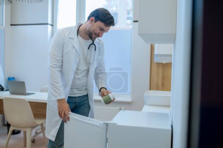 Téléchargez les photos : Jeune travailleur de la santé vêtu d'une blouse de laboratoire plaçant la bouteille de drogue au réfrigérateur - en image libre de droit