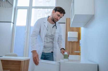 Téléchargez les photos : Jeune médecin concentré en blouse de laboratoire regardant dans le réfrigérateur ouvert de son bureau - en image libre de droit