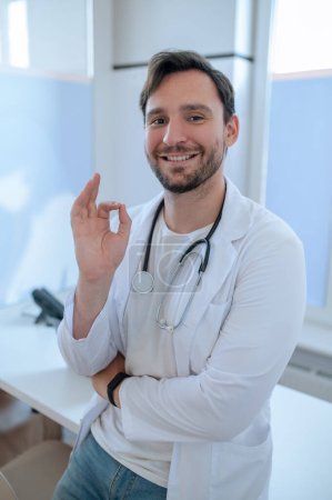 Téléchargez les photos : Médecin amical souriant en blouse de laboratoire appuyé sur le bureau et faisant le geste OK - en image libre de droit