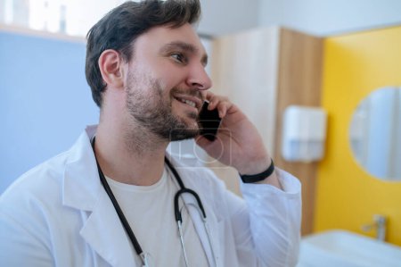 Téléchargez les photos : Souriant jeune médecin heureux en blouse de laboratoire faisant un appel téléphonique dans son bureau - en image libre de droit