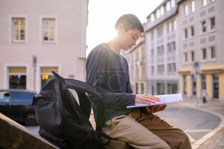 Photo for Serious concentrated adolescent boy sitting outdoors while flipping through the pages of a textbook - Royalty Free Image