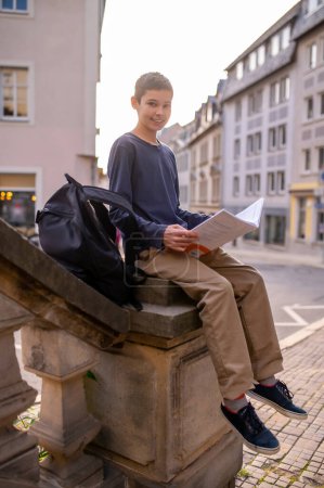 Photo for Smiling adolescent boy with an open book in his hands sitting on the balustrade and looking ahead - Royalty Free Image