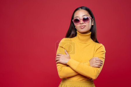 Foto de Retrato en la cintura de una chica tranquila y confiada en gafas de moda posando para la cámara sobre el fondo rojo - Imagen libre de derechos