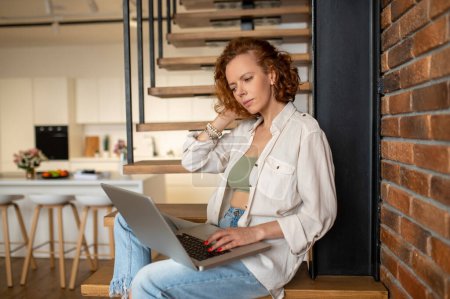 Téléchargez les photos : Travailler à la maison. Jeune travailleuse indépendante assise dans les escaliers avec un ordinateur portable - en image libre de droit