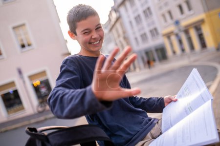 Téléchargez les photos : Adolescent riant assis avec un manuel ouvert à l'extérieur couvrant l'objectif de la caméra avec la paume de sa main - en image libre de droit
