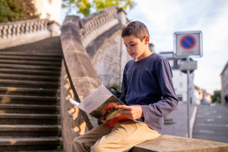Photo for Calm concentrated teenager sitting on the concrete balustrade with an open textbook in the hands - Royalty Free Image