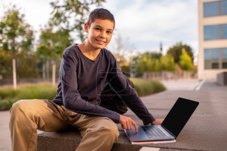 Sonriendo lindo adolescente sentado afuera tocando el touchpad en su computadora portátil y mirando antes que él