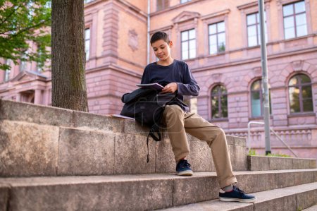 Photo for Smiling adolescent boy seated on the concrete stairs looking at a book in his hands - Royalty Free Image
