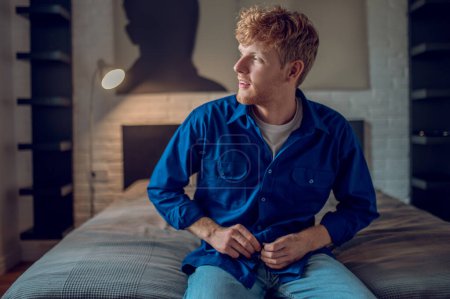 Photo for At home. A young man in dark-blue shirt sitting on the bed - Royalty Free Image