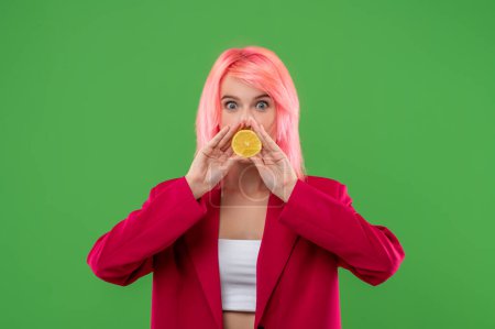 Photo for Waist-up portrait of a mischievous girl with pink hair posing with half of lemon against the green background - Royalty Free Image