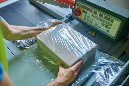 Téléchargez les photos : Photo recadrée d'un employé de l'entrepôt enveloppant la boîte en carton avec la machine d'emballage de film étirable - en image libre de droit