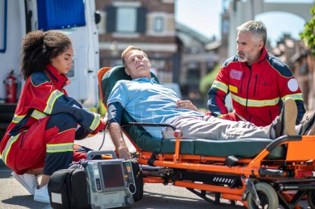 Téléchargez les photos : Un ambulancier spécialisé féminin enroule le brassard de pression artérielle autour du bras de la patiente supervisé par son collègue sérieux - en image libre de droit