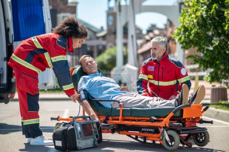 Téléchargez les photos : Médecin ambulancier sérieux fixant le brassard de pression artérielle sur le bras de la patiente en présence de son collègue - en image libre de droit