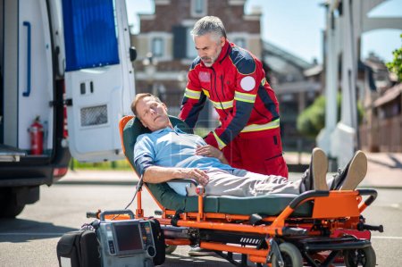 Téléchargez les photos : Un ambulancier sérieux penché sur un homme inconscient allongé sur le brancard est passé à l'ECG. - en image libre de droit