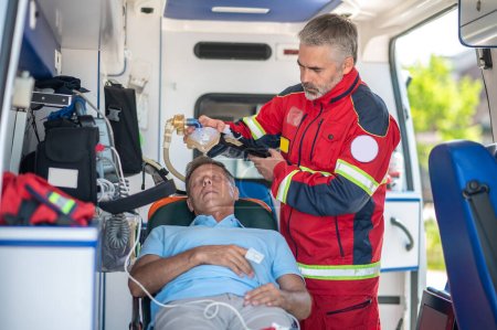 Téléchargez les photos : Un ambulancier paramédical sérieux pose le masque à oxygène sur le visage d'un homme inconscient allongé sur la civière - en image libre de droit
