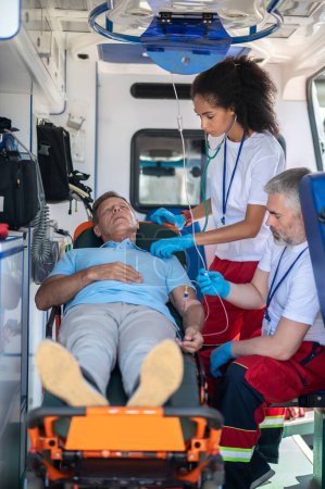 Téléchargez les photos : L'ambulancier écoute le cœur de la patiente avec le stéthoscope tandis que son collègue lui donne le goutte-à-goutte intraveineux - en image libre de droit