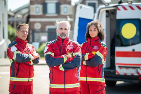 Photo for Team of serious paramedics dressed in the red uniforms standing outdoors beside the ambulance van - Royalty Free Image