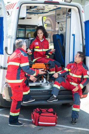 Photo for Smiling young female paramedic sitting on the ambulance gurney while discussing something with her colleagues - Royalty Free Image