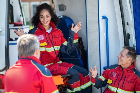 Photo for Cheerful female paramedic seated on the ambulance gurney having a lively conversation with her colleagues - Royalty Free Image
