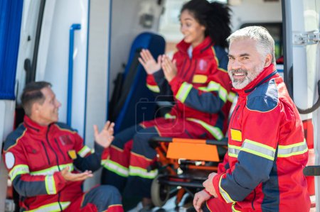 Sonriente alegre médico barbudo de pelo gris ambulancia de pie junto a sus colegas involucrados en una animada conversación