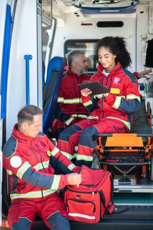 Téléchargez les photos : Joyeuse ambulancière paramédicale assise sur la civière de l'ambulance à l'aide de sa tablette pendant que ses collègues concentrés se préparent pour le travail - en image libre de droit