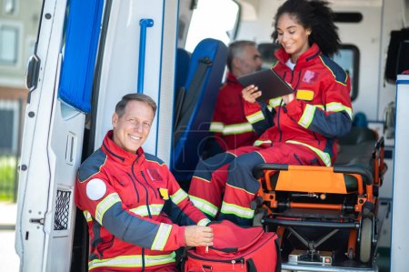 Téléchargez les photos : Amoureux ambulancier d'âge moyen et ses collègues en uniforme rouge se préparent pour leur quart de travail - en image libre de droit