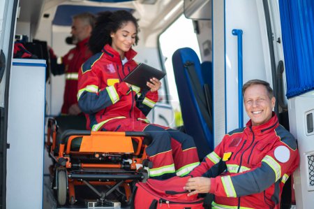 Téléchargez les photos : Joyeux médecin ambulancier d'âge moyen et ses collègues en uniformes rouges se préparant pour leur quart de travail - en image libre de droit