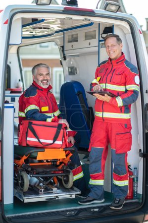 Smiling paramedic with a tablet computer standing beside his cheerful colleague seated on the ambulance gurney with a medical bag
