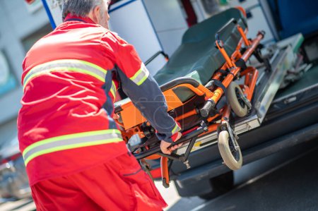 Photo for Back view of a gray-haired paramedical worker pulling the gurney out of the ambulance van - Royalty Free Image