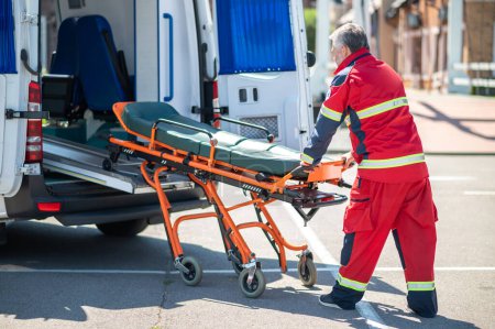 Foto de Trabajador paramédico vestido con el uniforme rojo sacando la camilla del vehículo EMS - Imagen libre de derechos