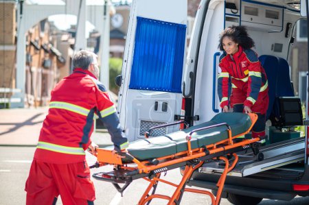 Photo for Young female paramedic pulling the gurney out of the ambulance van assisted by her colleague - Royalty Free Image