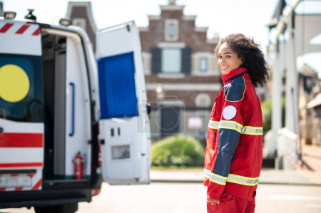 Photo for Smiling happy young female paramedic in the uniform standing in front of an EMS vehicle - Royalty Free Image