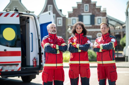 Photo for Pleased female paramedic and her colleagues in the red uniforms standing beside the EMS vehicle - Royalty Free Image