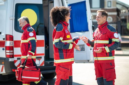 Téléchargez les photos : Des ambulanciers joyeux discutent à l'extérieur pendant que leur collègue avec le sac EMS passe devant eux - en image libre de droit