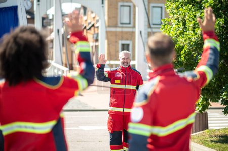 Téléchargez les photos : Ambulancier barbu aux cheveux gris joyeux dans l'uniforme rouge agitant deux collègues dans la rue - en image libre de droit