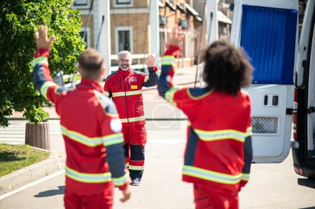 Téléchargez les photos : Joyeux médecin ambulancier mature en uniforme rouge agitant ses collègues dans la rue - en image libre de droit