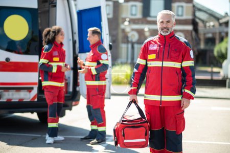 Photo for Calm mature gray-haired bearded paramedic posing for the camera with his colleagues on the background - Royalty Free Image